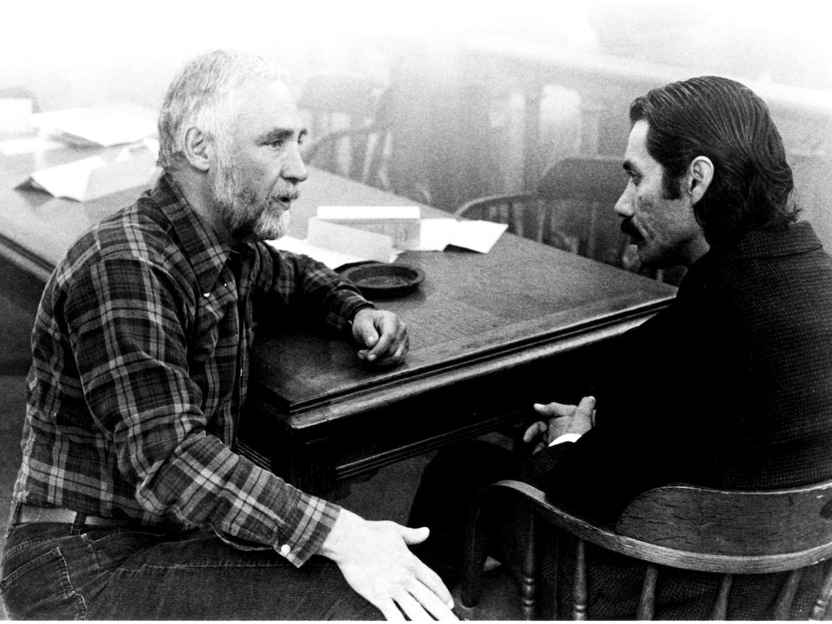 A bearded man in a plaid shirt and a mustached man in a dark jacket talk across a wooden table strewn with papers, a black-and-white still from The Ballad of Gregorio Cortez