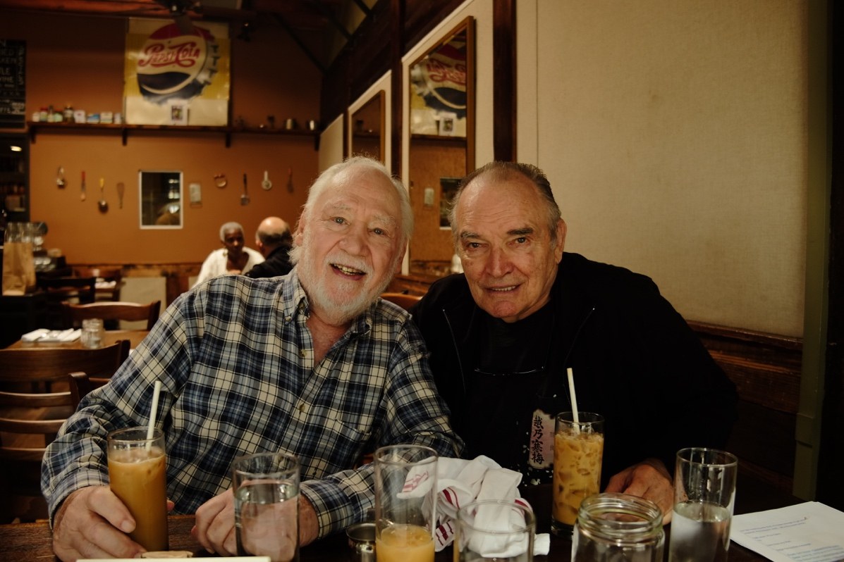 Two older men smile at a restaurant table over iced drinks, one bearded in a plaid shirt, the other in black