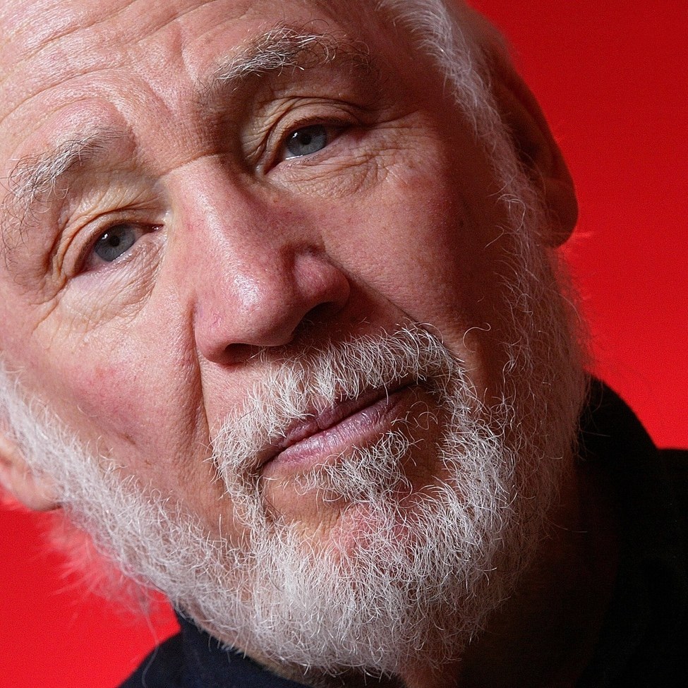 Close-up portrait of Robert M. Young, white-bearded, against a red background
